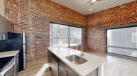 Located in the heart of Richmond, Virginia, this 2-bedroom, 1-bathroom residence masterfully combines historic industrial character with contemporary design. Exposed original brick walls and reclaimed timber columns serve as striking architectural focal points throughout, contrasting beautifully with clean white walls and light wood-look flooring that runs seamlessly across the entire floor.

The open kitchen and living area is a standout feature, showcasing a center island with quartz countertops and a stainless steel sink, dark wood cabinetry, and a full suite of stainless steel appliances including a refrigerator, electric range, and built-in microwave. A large sliding window floods the kitchen with natural light while framing views of the surrounding Richmond cityscape.

Both bedrooms offer soaring ceilings, generous natural light, and dedicated closet space. One bedroom features expansive windows set within the original brick facade, while the other highlights an exposed reclaimed timber column that adds warmth and historic depth. Two closets provide practical storage throughout the unit.

The bathroom is finished with a clean, functional aesthetic, featuring a full tub-shower combination. Ceiling fans in key living areas and light-toned flooring throughout complete this thoughtfully designed urban residence.