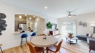 This single-floor residence offers a thoughtfully designed open-concept layout that seamlessly connects the living, dining, and kitchen areas. Light wood-style flooring flows throughout the main living spaces, complemented by neutral walls that create a bright and welcoming ambiance. The living room is anchored by a ceiling fan, large sliding glass doors that invite an abundance of natural light, and direct access to an outdoor area. The adjacent dining space features a round table and opens naturally into the kitchen through a distinctive arched passageway. The kitchen is well-equipped with stainless steel appliances, including a refrigerator, range, microwave, and dishwasher, all framed by contemporary wood-toned cabinetry and granite-style countertops. A breakfast bar with seating provides an additional casual dining option. The bedroom is a restful retreat with plush carpeting, generous natural light from large windows, and ample wall space. The bathroom is finished with matching wood-toned cabinetry, a granite-top vanity, wood-style flooring, and a full soaking tub with shower. Recessed lighting and ceiling fans throughout enhance both functionality and comfort across the entire space.