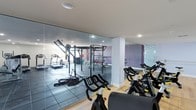 This fitness facility is thoughtfully organized into two dedicated workout zones, divided by a striking floor-to-ceiling glass partition that maintains visual openness while functionally separating the spaces. The cycling and cardio studio features warm hardwood flooring and houses a row of professional-grade stationary bikes, complemented by wall-mounted mirrors that run the length of the room, amplifying the sense of space. Subtle purple and lavender accent walls add a modern, energetic touch, while a ballet-style barre mounted along one wall suggests the space doubles as a group fitness or dance studio. Recessed lighting provides consistent, even illumination throughout. The adjacent strength training area transitions to durable dark rubber flooring, purpose-built for heavy equipment. It is outfitted with a multi-functional power rack, cable machines, treadmills, elliptical trainers, and battle ropes, catering to a full range of strength and cardio training needs. The glass partition connecting both zones allows users in each area to maintain sightlines across the facility, creating a cohesive and connected environment. The clean white ceilings and modern finishes throughout give the facility a polished, professional aesthetic.