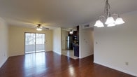 This single-floor residence is designed with an open-concept layout that connects the living area to the kitchen, creating a cohesive and functional flow throughout the space. Rich hardwood flooring extends across the main living area, adding warmth and a classic aesthetic, while the kitchen transitions to tile flooring. Crown molding lines the ceiling throughout, lending a polished finishing detail to the interior.

The living area is generously proportioned and bathed in natural light, courtesy of a large sliding glass door that opens to an outdoor space and offers views of the exterior. A ceiling fan with a light fixture is installed overhead, complementing an elegant chandelier near the dining area that adds a decorative touch.

The kitchen is partially open to the main living space, separated by a partial wall that maintains a sense of openness. It features dark cabinetry, a stainless steel refrigerator, and additional appliances, providing a well-equipped cooking environment. A door off the kitchen area leads to what appears to be an entry or utility space. Ceiling fans in both the living and kitchen areas ensure comfortable airflow throughout the home.