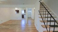 This two-floor residence offers a clean, functional layout with a bright and open feel throughout. The main level features light-toned hardwood-style flooring that flows seamlessly across the living area, entryway, and into the kitchen, creating a cohesive and airy atmosphere. The living space is generously proportioned, providing ample room for comfortable furnishing arrangements.

The kitchen is equipped with dark wood cabinetry, a white refrigerator, and a dishwasher, with a window above the sink that invites natural light into the space. A ceiling fan is present, adding to the room's practicality. The front entry is defined by a classic white door with decorative glass insets, offering a welcoming point of arrival.

A standout architectural feature is the open staircase, which showcases natural wood treads paired with a sleek black metal railing — a modern design element that adds visual interest and contrast against the home's predominantly white walls. The crisp white interior walls throughout lend a fresh, neutral canvas well-suited for personalization. Together, these elements combine to create a practical yet stylish home with a clean, updated aesthetic across both levels.