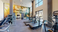 This expansive fitness center is designed to inspire and support a comprehensive workout experience. The space is defined by dramatic double-height ceilings and floor-to-ceiling windows that flood the interior with natural light while offering views of the surrounding outdoor environment. A bold, large-scale motivational mural anchors the main workout area, setting an energetic tone throughout.

The main floor is generously outfitted with a full row of commercial-grade treadmills, elliptical trainers, stair climbers, stationary bikes, and rowing machines to cover all cardio needs. A dedicated strength training section features a multi-use cable machine, adjustable benches, a complete dumbbell rack, and a punching bag setup. A water fountain station with tiled accent wall detailing provides convenient hydration access.

A separate studio room offers a mirrored wall with a ballet barre, stability balls, foam rollers, kettlebells, and a wall-mounted TV, making it ideal for yoga, barre, stretching, and group fitness activities. The studio's high ceilings and herringbone-patterned carpet flooring carry through the same refined aesthetic found throughout the facility.

Throughout the space, warm pendant lighting, neutral-toned walls, and a cohesive herringbone carpet design contribute to an upscale yet functional ambiance built for serious fitness pursuits.