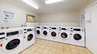 Located at 101 Hutson Dr in Summerville, South Carolina, this single-floor community laundry facility is designed to provide a clean, functional, and well-organized space for residents. The room features light-toned tile flooring throughout, bright overhead fluorescent lighting, and neutral painted walls that give the space an open and tidy feel. Crown molding along the ceiling adds a subtle finishing touch to the interior.

Along one wall, a row of numbered front-load washing machines is neatly arranged side by side, each equipped with digital controls for ease of use. On the opposite side, a bank of stacked dryers — also individually numbered — maximizes the available space while accommodating high-volume laundry needs. Natural light filters in through a window with blinds near the dryer section, complementing the overhead lighting.

A dedicated folding and waiting area occupies one end of the room, complete with a fold-out table for sorting and folding laundry and a pair of seating chairs for convenience. Informational signage and laundry tips are posted on the walls, and a digital payment kiosk is mounted nearby for easy access. A fire extinguisher is prominently displayed, and rubber entry mats are positioned at key areas throughout the room. A community bulletin board rounds out this practical and well-maintained shared facility.
