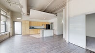 This single-floor loft-style unit showcases a thoughtfully designed open-concept layout that seamlessly blends industrial character with modern finishes. Soaring ceilings with exposed beams, ductwork, and structural elements lend an airy, warehouse-inspired aesthetic throughout the main living area, which is bathed in natural light from a generous bank of large windows overlooking greenery outside.

The kitchen is well-appointed with light maple cabinetry offering ample storage, dark granite-style countertops, black appliances including a full-size refrigerator and dishwasher, a built-in cooktop on the central island, and tile flooring that transitions smoothly to the light hardwood-style floors of the living area. A partial ceiling drop above the kitchen zone creates a subtle separation between cooking and living spaces.

Two bathrooms serve the unit. One features a clean, minimalist design with a pedestal sink, a standalone shower stall, and tile flooring. The second offers a more refined finish with a dual-sink vanity, warm wood cabinetry, a wide mirror with overhead lighting, and tile floors. A dedicated laundry area includes a stacked washer and dryer for added convenience. The space presents a versatile, light-filled environment well-suited for a variety of living arrangements.