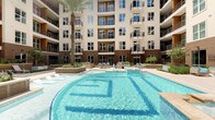 This residential community showcases two distinct shared amenity spaces designed to enhance everyday living. The centerpiece is a stunning resort-style outdoor pool, set within a landscaped courtyard framed by the building's multi-story facade. The pool features a freeform design with crystal-clear blue water, decorative tile detailing along the pool floor, and surrounding deck areas furnished with lounge seating. Tall palm trees and planters add a tropical ambiance, while the building's balconies overlook the space, creating an enclosed and private atmosphere. The area also includes a spa or wading zone, completing the resort-inspired experience.

The interior mail room offers a clean, functional design with a contemporary aesthetic. Painted in a soft teal-blue palette with light tile flooring, the room is lined with rows of individual bronze mailboxes mounted along both sides. A centrally positioned parcel and sorting counter provides convenient mail handling. Large floor-to-ceiling windows at one end flood the space with natural light and offer views of surrounding greenery. Framed abstract artwork adorns the walls, adding a touch of sophistication. Recessed lighting complements the natural light, creating a bright and welcoming environment for residents.