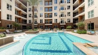This residential community showcases two distinct shared amenity spaces designed to enhance everyday living. The centerpiece is a stunning resort-style outdoor pool, set within a landscaped courtyard framed by the building's multi-story facade. The pool features a freeform design with crystal-clear blue water, decorative tile detailing along the pool floor, and surrounding deck areas furnished with lounge seating. Tall palm trees and planters add a tropical ambiance, while the building's balconies overlook the space, creating an enclosed and private atmosphere. The area also includes a spa or wading zone, completing the resort-inspired experience.

The interior mail room offers a clean, functional design with a contemporary aesthetic. Painted in a soft teal-blue palette with light tile flooring, the room is lined with rows of individual bronze mailboxes mounted along both sides. A centrally positioned parcel and sorting counter provides convenient mail handling. Large floor-to-ceiling windows at one end flood the space with natural light and offer views of surrounding greenery. Framed abstract artwork adorns the walls, adding a touch of sophistication. Recessed lighting complements the natural light, creating a bright and welcoming environment for residents.