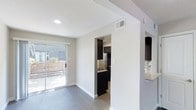 This two-floor space showcases a clean, modern design with a neutral color palette throughout. The main living area features wood-style flooring in a cool gray tone that complements the crisp white walls, creating a bright and contemporary atmosphere. Recessed lighting overhead adds to the well-lit environment, which is further enhanced by abundant natural light streaming through a full-width sliding glass door. The slider opens to an outdoor area enclosed by a wooden fence, offering a private exterior space.

An open passageway connects the living area to the kitchen, which is outfitted with dark espresso-toned cabinetry that provides a striking contrast against the lighter surroundings. The kitchen includes stainless steel appliances, including a refrigerator and range, along with a tiled backsplash that adds a decorative touch. The cabinetry extends to both upper and lower configurations, offering ample storage. Structural beam details are visible along the ceiling, adding an architectural element to the space. The layout promotes a functional flow between the living and kitchen areas, making the space practical for everyday use across both floors.