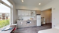 This thoughtfully designed single-floor studio offers a clean, contemporary aesthetic throughout. The open-plan layout seamlessly integrates the living, sleeping, and kitchen areas, creating a cohesive and efficient use of space. Wood-look plank flooring runs throughout, lending warmth and visual continuity to every area.

The kitchenette is well-appointed with two-tone cabinetry in white and taupe, a smooth cooktop, built-in microwave, single-basin sink, and a full-size refrigerator. Open shelving provides convenient additional storage. Recessed lighting illuminates the kitchen workspace effectively.

The sleeping area benefits from a large window that invites generous natural light and offers views of the surrounding residential exterior. A ceiling fan adds comfort and airflow, while the bright white walls contribute to an open, airy atmosphere. A compact dining setup nestled against the window creates a functional space for meals with a pleasant outdoor outlook.

The bathroom features wood-look flooring consistent with the rest of the unit, a combined shower and bathtub, toilet, and a vanity with storage. Clean white finishes keep the space feeling fresh and bright.

The entry foyer provides a defined transition into the unit, complete with a secure door. Overall, the space offers a practical, modern living environment with a cohesive and neutral design palette.