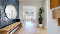 This thoughtfully designed single-floor space exudes a modern, sophisticated aesthetic from the moment you enter. The entry area makes an immediate impression with a striking navy blue vertical tile feature wall, a live-edge wood console, and a round mirror — setting a refined tone that carries throughout the space. Underfoot, light herringbone-patterned hardwood flooring ties each area together seamlessly, adding warmth and visual interest.

The open layout flows effortlessly between a welcoming reception-style lounge and a dedicated meeting or dining area. The lounge features a sleek gray upholstered bench seat paired with gold-accented side tables resting on marble bases, alongside lush potted plants that bring life and texture to the bright, white-walled interior. Large arched windows dressed with roller shades allow generous natural light to fill the space while framing views of the surrounding outdoor environment.

The dining area is anchored by a substantial live-edge wood table surrounded by plush gray upholstered chairs, beneath a bold sputnik-style chandelier. Abstract wall art in muted blue tones complements the overall color palette. French doors at the rear of the space provide additional natural light and a sense of openness, completing this polished, design-forward interior.