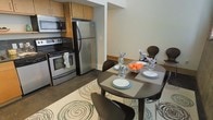 This two-floor space is designed with a practical and modern aesthetic that balances functionality with contemporary style. The kitchen is equipped with stainless steel appliances, including a full-size refrigerator, an over-the-range microwave, and an electric range, all complemented by warm wood-toned cabinetry that provides ample storage. The dark flooring throughout the kitchen area adds a sleek, grounded contrast to the light-toned walls.

Adjacent to the kitchen, the dining area flows naturally from the cooking space, creating an open and connected layout ideal for everyday living and entertaining. A dark wood dining table anchored on a patterned area rug with circular motifs seats multiple guests comfortably, adding a casual yet refined touch to the space. The neutral wall tones and clean lines throughout contribute to a bright, airy ambiance.

The two-floor layout suggests additional living, sleeping, or utility spaces beyond what is immediately visible, offering flexibility and room for a variety of living arrangements. Overall, the space presents a cohesive, move-in-ready environment with a clean, urban character.