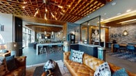 This single-floor amenity space delivers a sophisticated industrial-modern aesthetic, blending warm natural materials with sleek contemporary finishes throughout. The centerpiece of the main lounge is a dramatic wood lattice ceiling punctuated by a striking sputnik chandelier, creating an eye-catching overhead feature that sets the tone for the entire space. Floor-to-ceiling windows flood the lounge with abundant natural light and offer views of the surrounding outdoor greenery, while tufted cognac leather sofas and layered textiles establish an inviting, upscale atmosphere.

A built-in coffee and refreshment bar serves as a social hub, positioned adjacent to a communal high-top counter with bar seating, ideal for casual gatherings or informal work sessions. Exposed brick walls, stone-tile surfaces, and wood-slat partitions add rich textural contrast throughout the space.

A dedicated games area features a full-size billiards table beneath a cluster of pendant lights, complemented by intimate bistro-style seating along a textured brick wall. A separate coworking zone, enclosed by floor-to-ceiling glass partitions, provides a quiet, focused environment with individual workstations and ample desk space.

Private booth seating alcoves with wood-paneled walls and built-in banquettes offer semi-secluded nooks for small meetings or relaxed conversation. A well-appointed business room rounds out the space, featuring a large wood conference table, task seating, and a bold graphic wall mural, making this a fully functional and visually dynamic communal environment.