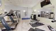 Located at 160 Chapel Street in Ottawa's Sandy Hill neighbourhood, this fitness center offers a well-equipped, single-floor exercise space designed to support a comprehensive workout routine. The room features light-toned tile flooring throughout and a dropped ceiling with recessed panel lighting, creating a clean and functional atmosphere.

The cardio zone is equipped with multiple treadmills, elliptical trainers, and a stationary cycle, positioned near a large window that allows natural light to fill the space and provides an outdoor view. A multi-station cable machine and a dedicated free weights area with a full dumbbell rack and adjustable bench cater to strength training needs. A large blue exercise mat provides a designated stretching and floor exercise zone.

Wall-mounted mirrors visually expand the room and allow users to monitor their form, while a wall-mounted television adds an entertainment option during workouts. Sanitation supplies, informational postings, and a first aid station are conveniently accessible along one wall, reflecting attention to safety and hygiene. A gray double door provides access to the corridor, ensuring easy entry and exit. The overall environment is bright, organized, and purpose-built to meet a wide range of fitness needs.