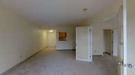 This single-floor residence offers a straightforward and functional layout designed for comfortable everyday living. The main living area is generously proportioned, featuring soft, neutral carpeting that adds warmth underfoot and smooth, light-toned walls that contribute to a bright and airy atmosphere. Ceiling-mounted light fixtures provide consistent illumination throughout the space.

A pass-through opening connects the living area to the kitchen, which is outfitted with warm wood-toned cabinetry and a countertop that offers ample workspace. This design element creates a sense of openness between the cooking and living spaces while maintaining a defined boundary.

A set of bifold closet doors provides accessible storage directly within the main living area, offering practical organization within easy reach. An additional doorway leads to a secondary space, suggesting a private room or utility area beyond.

The entryway, visible in the background, features a white panel door and is well-lit, creating a welcoming point of arrival. The overall interior palette — composed of soft whites, beige carpeting, and natural wood accents — lends the space a calm, neutral character well-suited to a variety of furnishing styles and personal tastes.