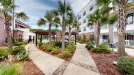 Located at 1813 Sir Tyler Dr in Wilmington, North Carolina, this outdoor amenity space offers a well-appointed, resort-style environment designed for relaxation and socializing. At the heart of the space is a generously sized swimming pool with clearly marked depth indicators and multiple entry points via stainless steel railings. The pool deck is finished in a textured aggregate surface and lined with cushioned lounge chairs in teal and gray tones, providing ample space for sunbathing.

Surrounding the pool, mature palm trees and manicured landscaping create a coastal ambiance, with mulched garden beds, low shrubs, and decorative plantings adding lush greenery throughout. Winding paver pathways connect various seating areas and lead to a series of wooden pergolas that offer shaded gathering spots.

A dedicated outdoor lounge area features Adirondack-style chairs positioned around a brick fire pit, ideal for cooler evenings. Mounted on an adjacent brick wall is a large flat-screen outdoor television, complemented by a sound bar, making it a functional entertainment hub. Additional seating, including picnic-style tables under a covered pergola, rounds out the space.

The surrounding multi-story building features a classic combination of warm brick and white siding, with balconies overlooking the courtyard, contributing to an enclosed, community-centered atmosphere.