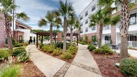 Located at 1813 Sir Tyler Dr in Wilmington, North Carolina, this outdoor amenity space offers a well-appointed, resort-style environment designed for relaxation and socializing. At the heart of the space is a generously sized swimming pool with clearly marked depth indicators and multiple entry points via stainless steel railings. The pool deck is finished in a textured aggregate surface and lined with cushioned lounge chairs in teal and gray tones, providing ample space for sunbathing.

Surrounding the pool, mature palm trees and manicured landscaping create a coastal ambiance, with mulched garden beds, low shrubs, and decorative plantings adding lush greenery throughout. Winding paver pathways connect various seating areas and lead to a series of wooden pergolas that offer shaded gathering spots.

A dedicated outdoor lounge area features Adirondack-style chairs positioned around a brick fire pit, ideal for cooler evenings. Mounted on an adjacent brick wall is a large flat-screen outdoor television, complemented by a sound bar, making it a functional entertainment hub. Additional seating, including picnic-style tables under a covered pergola, rounds out the space.

The surrounding multi-story building features a classic combination of warm brick and white siding, with balconies overlooking the courtyard, contributing to an enclosed, community-centered atmosphere.