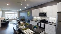 This contemporary single-floor residence is designed with an open-concept layout that seamlessly connects the kitchen and living areas, creating a cohesive and functional living environment. The kitchen is a standout feature, showcasing crisp white cabinetry that extends to the ceiling, striking dark granite countertops, and a full suite of stainless steel appliances, including a range and over-the-range microwave. A mosaic tile backsplash adds a refined textural element, complementing the kitchen's clean, modern aesthetic. The expansive island with bar seating serves as both a practical workspace and an inviting gathering spot. Track lighting and recessed ceiling lights provide layered illumination throughout. Bold gray accent walls add depth and contrast against the white cabinetry, while vibrant artwork infuses a pop of color and personality into the space. The living area beyond the kitchen is bathed in natural light from large windows, offering views of the surrounding cityscape. The overall ambiance is sophisticated and urban, blending stylish design elements with thoughtful functionality to create a welcoming and well-appointed living space.