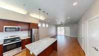 This two-story townhouse in Cedar Park, TX offers a well-designed layout spread across two floors. The main level features an open-concept kitchen and living area finished with warm hardwood-style flooring throughout. The kitchen is equipped with rich dark cabinetry, stainless steel appliances including a refrigerator, range, microwave, and dishwasher, and a central granite-topped island illuminated by elegant pendant lighting. The living area extends naturally from the kitchen, enhanced by a ceiling fan and a sliding glass door that invites natural light and outdoor views. Upstairs, two bedrooms are finished with plush carpet and neutral tones. The primary bedroom boasts soaring vaulted ceilings, a large sliding glass door opening to a private balcony with outdoor views, and access to a dedicated walk-in closet with built-in shelving. The bathroom features a dual-sink granite vanity with a large mirror and wood-toned cabinetry, maintaining design continuity throughout the home. A second bedroom with hardwood-style flooring provides additional flexible space. A staircase with wooden treads connects both floors, completing this functional and stylish townhouse.