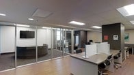 This contemporary single-floor office space combines open-plan functionality with thoughtfully designed private areas, creating a professional and productive environment. The layout features a welcoming reception or workstation area finished with a sleek, light-toned countertop surface, complemented by warm hardwood-style flooring that runs throughout the main corridor and open zones.

Floor-to-ceiling glass partition walls with slim aluminum framing divide the space into distinct enclosed rooms while maintaining a sense of openness and transparency. These glass-walled offices and meeting rooms are furnished with comfortable seating and allow natural light to filter through, brightening the entire floor. Interior windows within some partitioned rooms further enhance the flow of light across the space.

The ceiling features a clean drop-tile design with flush and recessed panel lighting, ensuring consistent, well-distributed illumination throughout. A darker accent wall adds depth and visual contrast to one section of the office, balancing the otherwise bright and neutral palette.

Workstation areas are thoughtfully arranged to support collaboration, while the enclosed glass rooms offer privacy for focused work or client meetings. The overall ambiance is professional, modern, and well-appointed — designed to accommodate a range of office functions within a cohesive and polished environment.