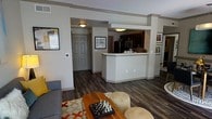 This single-floor residence features an open-concept design that integrates the living area and kitchen into one cohesive space. Dark hardwood-style flooring runs throughout, providing a rich contrast against the neutral gray walls and white trim. The living area is comfortably proportioned, accommodating a sofa, coffee table, and accent seating, with framed artwork adding visual interest to the walls.

A half-wall peninsula separates the kitchen from the main living space while maintaining an open sightline. The kitchen is equipped with dark cabinetry, stainless steel appliances including a refrigerator and microwave, and offers functional counter space for meal preparation. Recessed ceiling fixtures and flush-mount lighting provide consistent illumination throughout the space.

An entryway is visible beyond the living area, featuring a gray paneled front door and a dedicated hallway that provides clear separation between the entry and main living zones. Crown molding and architectural column details add a subtle refined quality to the overall design. The layout is practical and well-organized, making efficient use of the available floor space.