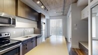 Located on the third floor at 10620 116 Street NW in Edmonton, this one-bedroom, one-bathroom unit showcases a bold industrial-modern aesthetic that makes a strong architectural statement. Exposed concrete ceilings with track lighting run throughout the main living area and kitchen, adding an urban edge complemented by warm hardwood-style flooring underfoot.

The kitchen is a standout feature, offering dark wood cabinetry, light quartz countertops, a generous central island, and a full suite of stainless steel appliances including a refrigerator, electric range, and over-the-range microwave. A stacked in-unit washer and dryer is also thoughtfully integrated into the kitchen area. The layout flows efficiently from the kitchen into the open living space, which benefits from a large floor-to-ceiling window dressed with vertical blinds, allowing ample natural light to fill the room.

A sliding glass partition separates the living area from the bedroom, which features its own window and ceiling fan, maintaining an airy feel. The hallways connect each space with clean sightlines throughout. The bathroom is finished with large-format white tiles, a soaking tub with shower, and consistent hardwood-style flooring that ties the unit together cohesively. This unit offers a refined, contemporary living experience in a compact yet well-designed footprint.