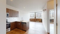 Located at 3715 Main Street in Philadelphia's Manayunk neighborhood, this one-bedroom, one-bathroom unit blends industrial loft character with contemporary design. The open-plan kitchen and living area serves as the heart of the home, where warm hardwood flooring flows throughout and expansive factory-style grid windows flood the space with natural light. Original exposed brick along the lower walls and a structural concrete column add authentic industrial charm, while a wall-mounted mini-split system ensures modern climate control.

The kitchen is well-appointed with rich walnut-toned cabinetry, a full suite of stainless steel appliances including a refrigerator, range, built-in microwave, and dishwasher, and a clean white quartz peninsula countertop that opens to the living area. The spacious bedroom continues the hardwood flooring and benefits from its own wall-mounted AC unit, offering a comfortable and quiet retreat. A dedicated closet provides practical storage.

The bathroom features a sleek gray vanity with white countertop, a large mirror, and industrial-style exposed pipe light fixture, complemented by dark textured flooring that reinforces the unit's loft aesthetic. The overall ambiance is bright, airy, and urban, with a distinctive character rooted in the building's industrial past.