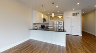 This thoughtfully designed single-floor residence offers a clean, modern aesthetic throughout, combining functionality with stylish finishes. The open-concept living and kitchen area serves as the heart of the space, anchored by a striking kitchen featuring white shaker cabinetry, dark countertops, a marble-style tile backsplash, stainless steel appliances, and elegant pendant lighting. A generous kitchen island provides additional prep space and a natural divide between the kitchen and the expansive living area. Rich hardwood flooring runs throughout the main living spaces, adding warmth and continuity to the overall design. A wall of large, multi-pane windows floods the living area with natural light, creating a bright and airy atmosphere. Recessed lighting is installed throughout, ensuring well-lit interiors at all times. The private rooms are finished with the same warm hardwood flooring, neutral walls, and clean white trim, offering versatile spaces adaptable to a variety of uses. An in-unit stacked washer and dryer adds everyday convenience. The full bathroom is finished with large-format neutral wall tiles, a soaking tub with shower, a white vanity, and polished chrome fixtures, delivering a clean and contemporary feel. Throughout the space, a neutral color palette and modern hardware selections create a cohesive and move-in-ready environment.