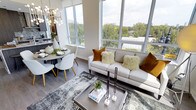 This stylish single-floor space is defined by its expansive floor-to-ceiling windows, which flood the interior with abundant natural light and frame sweeping views of a lush, tree-lined urban landscape. The open-concept layout connects the living and dining areas in a cohesive, functional arrangement that maximizes the sense of space.

The living area is anchored by a sleek, light-toned sofa adorned with richly textured amber and ivory throw pillows, complemented by a contemporary metallic coffee table and a patterned area rug. Tall ceilings enhance the airy atmosphere, while an eye-catching geometric chandelier adds a sculptural focal point overhead.

The dining area features a minimalist round table paired with modern white chairs accented with gold-toned legs, creating an inviting spot for meals. Decorative accents, including curated tableware and greenery, add warmth to the otherwise clean aesthetic.

Hardwood-style flooring runs throughout, tying the spaces together cohesively. The overall ambiance is sophisticated yet welcoming, blending modern design elements with warm tones to create a bright, contemporary living environment.