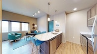 This single-floor space showcases a thoughtfully designed open-concept layout that connects the kitchen and living area in a cohesive, functional arrangement. The kitchen serves as a central feature, anchored by a curved peninsula with a striking white marble-veined countertop that provides both prep space and casual seating for multiple bar-height chairs in a bold teal finish. Dark cabinetry beneath the peninsula offers generous storage, while a stainless steel sink and dishwasher add modern utility. Overhead, two sleek pendant lights illuminate the island, complementing a track lighting system that runs across the ceiling toward the living area.

The living space flows naturally from the kitchen, featuring warm wood-look flooring throughout that ties the zones together. Neutral wall tones in the main area contrast with an accent wall in a deep taupe, adding visual depth. A window brings in natural light, enhancing the airy feel of the space. A teal entry door adds a vibrant pop of color that echoes the seating accents. Overall, the space balances contemporary finishes with functional design.