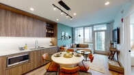 This thoughtfully designed single-floor residence offers a seamless open-concept layout that integrates the kitchen, dining, and living areas into one cohesive space. The kitchen is outfitted with wood-grain cabinetry extending to the ceiling, open shelving, a gas cooktop, built-in oven, and light stone countertops paired with a white tile backsplash. A sleek track lighting system overhead illuminates the space efficiently. The dining area accommodates a round white table surrounded by warm amber leather chairs, positioned between the kitchen and living room for easy flow.

The living room is anchored by a large sectional sofa set against a muted teal accent wall, with floor-to-ceiling windows and a glass door offering expansive, elevated city views and abundant natural light. Light wood flooring runs throughout the main living areas.

The primary bedroom continues the floor-to-ceiling window theme, featuring a sage green accent wall and sweeping urban views. Two bathrooms serve the unit: one with a glass-enclosed walk-in shower, wood vanity, terrazzo flooring, and a lit mirror with open shelving; the other with a soaking tub, wood cabinetry, and matching terrazzo floors.