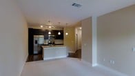 This single-floor space showcases an open-concept layout that connects the kitchen and living area in a cohesive, functional design. The kitchen is appointed with dark espresso cabinetry that contrasts sharply against light-colored walls, creating a bold visual dynamic. Stainless steel appliances, including a French door refrigerator with ice and water dispenser and an over-the-range microwave, provide modern utility. A central island with a light stone countertop offers additional workspace and casual seating, anchored by a track lighting system and elegant pendant lights suspended from the ceiling that illuminate the space effectively. A tile backsplash adds a refined detail to the cooking area. The flooring transitions from rich dark hardwood in the kitchen zone to plush carpet in the living area, delineating the two spaces while maintaining an open feel. A hallway visible in the background leads to additional rooms, suggesting a well-organized floor plan. The overall ambiance is contemporary and practical, with neutral warm tones throughout the walls and trim contributing to a welcoming, move-in-ready atmosphere.