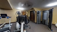 This single-floor fitness room offers a practical and comfortable environment for exercise and physical wellness. The space is finished with wall-to-wall carpeting and warm yellow-toned walls that create an inviting atmosphere. Overhead, a ceiling fan provides air circulation alongside a flush-mounted light fixture, ensuring the room remains well-ventilated and adequately lit during workouts.

The room is equipped with a selection of exercise machines, including an elliptical trainer prominently positioned toward the front of the space, ideal for low-impact cardio workouts. To the right, a multi-function cable resistance training machine offers a range of strength and toning exercises. Additional equipment is visible along the walls, providing users with varied workout options.

Architectural details include a sloped ceiling section in one corner, adding visual character to the room, along with classic wood-framed doors that lend a traditional aesthetic. A mirrored section near the entrance enhances the sense of depth and allows users to monitor their form during exercise.

Overall, this fitness room is a well-organized, fully functional space that balances practicality with a warm, approachable design, making it suitable for a range of fitness routines in a welcoming setting.