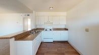 This single-story space offers a clean, functional layout designed with everyday comfort in mind. The kitchen serves as a standout feature, showcasing crisp white cabinetry that extends from base to upper wall level, providing generous storage throughout. Dark granite-style countertops offer a rich contrast against the bright white surfaces, while a double-basin sink adds practical utility. The kitchen is equipped with a white electric range and is complemented by a ventilation hood above. Warm wood-look flooring runs throughout the kitchen area, adding a welcoming and grounded aesthetic to the space. The bright white walls and flush ceiling light fixture keep the atmosphere clean and well-lit. A glimpse into the adjoining areas reveals additional rooms accessible via a hallway, suggesting a connected and thoughtfully arranged floor plan. Natural light appears to filter in from nearby spaces, contributing to the overall open and airy feel of the interior. The space presents a straightforward, move-in-ready environment with a neutral palette that lends itself well to a variety of interior styles and furnishing choices.