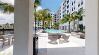 This expansive outdoor amenity area is designed with a resort-inspired aesthetic, centered around a large, shimmering pool flanked by tall swaying palm trees and lined with lounge chairs and market umbrellas. The surrounding hardscape features light-toned stone pavers that extend throughout the space, lending a clean, cohesive look. Adjacent to the pool, a sleek fire pit with stylish woven lounge chairs provides an inviting gathering spot set against a backdrop of modern, white multi-story buildings.

A covered outdoor kitchen and dining area offers a well-appointed space for entertaining, complete with a stainless steel gas grill set into granite countertops, a utility sink, and additional prep space. Dining tables and chairs are arranged beneath the covered structure, which features soaring ceilings with a ceiling fan and a wall-mounted television, making it functional in any weather. The area flows seamlessly between shaded and open-air zones.

The perimeter of the property incorporates lush tropical landscaping, including native grasses and mature palms, bordered by a black wrought-iron fence that opens to serene waterfront views beyond. The overall ambiance is bright, airy, and upscale, reflecting a South Florida lifestyle with thoughtful design throughout.