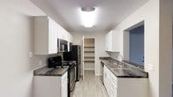 This single-floor space centers around a thoughtfully designed galley-style kitchen that serves as its standout feature. The kitchen is laid out with efficiency in mind, offering parallel runs of cabinetry on both sides that maximize storage and workflow. White shaker-style upper and lower cabinets line the walls, providing ample storage and contributing to a clean, bright aesthetic. The countertops feature a dark, veined stone surface that creates a striking contrast against the white cabinetry.

The kitchen is equipped with a full suite of stainless steel appliances, including a refrigerator, built-in microwave, and a black electric range with stovetop. A stainless steel double sink with a modern arc-neck faucet is positioned along one counter run. Overhead, a flush-mount ceiling light provides consistent illumination throughout the space.

A notable functional element is the open walk-in pantry located at the far end of the kitchen, fitted with multiple built-in shelves ideal for organized storage. The light wood-toned flooring runs throughout, complementing the overall neutral color palette and reinforcing the space's clean, modern character.