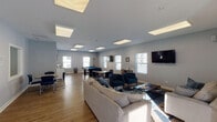 Located at 521 Fuller Ave in Big Rapids, Michigan, this single-floor community amenity center is designed to offer residents a comfortable and versatile gathering space. The layout encompasses several distinct areas that flow seamlessly together, all finished with light gray walls, wood-look flooring, and flush panel ceiling lights that create a bright, modern atmosphere throughout.

The main lounge and game room is the centerpiece of the facility — a generously sized open area illuminated by multiple large skylight-style ceiling panels and flanked by white-shuttered windows that invite natural light. This space features a full-size pool table, casual seating arrangements with sofas and blue accent chairs, wall-mounted televisions, and dining-height tables for group activities or socializing.

A dedicated private lounge offers a more intimate setting, furnished with a loveseat, armchair, coffee table, and its own wall-mounted TV — ideal for small group gatherings or quiet relaxation.

The fitness center is a standout feature, divided across two connected rooms. Equipment includes treadmills, an elliptical, a rowing machine, a stationary bike, a full dumbbell rack, and open rubber-matted floor space for stretching or floor exercises. Wall-mounted TVs and ample natural light enhance the workout experience.

Together, these amenities provide a functional and inviting community hub suited for recreation, fitness, and social connection.