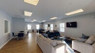 Located at 521 Fuller Ave in Big Rapids, Michigan, this single-floor community amenity center is designed to offer residents a comfortable and versatile gathering space. The layout encompasses several distinct areas that flow seamlessly together, all finished with light gray walls, wood-look flooring, and flush panel ceiling lights that create a bright, modern atmosphere throughout.

The main lounge and game room is the centerpiece of the facility — a generously sized open area illuminated by multiple large skylight-style ceiling panels and flanked by white-shuttered windows that invite natural light. This space features a full-size pool table, casual seating arrangements with sofas and blue accent chairs, wall-mounted televisions, and dining-height tables for group activities or socializing.

A dedicated private lounge offers a more intimate setting, furnished with a loveseat, armchair, coffee table, and its own wall-mounted TV — ideal for small group gatherings or quiet relaxation.

The fitness center is a standout feature, divided across two connected rooms. Equipment includes treadmills, an elliptical, a rowing machine, a stationary bike, a full dumbbell rack, and open rubber-matted floor space for stretching or floor exercises. Wall-mounted TVs and ample natural light enhance the workout experience.

Together, these amenities provide a functional and inviting community hub suited for recreation, fitness, and social connection.
