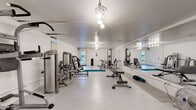 Located at 1800 Lakewood Dr in Phenix City, Alabama, this fitness center offers a comprehensive, single-floor exercise facility designed to support a full range of workout routines. The space is finished with clean white walls and a smooth light-colored floor, creating a bright and open atmosphere that encourages an energetic training environment.

The gym is generously equipped with a variety of Fitco strength-training machines, including cable stations, leg press units, and multi-function resistance equipment, providing options for both upper and lower body conditioning. A dedicated free weights and benching area complements the strength equipment, while a separate cardio zone houses treadmills, elliptical trainers, an inversion table, and a stationary bike, ensuring a well-rounded fitness experience.

A standout feature of the main training area is the full-length wall mirror, which spans a significant portion of one side of the room, visually expanding the space and allowing users to monitor their form during workouts. Overhead pendant lighting illuminates the entire floor evenly. A teal exercise mat and foam rollers are available for stretching and flexibility work. A fire extinguisher and posted rules signage reflect attention to safety. Natural light filters in through windows and a door at one end, offering views of the surrounding greenery.
