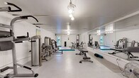 Located at 1800 Lakewood Dr in Phenix City, Alabama, this fitness center offers a comprehensive, single-floor exercise facility designed to support a full range of workout routines. The space is finished with clean white walls and a smooth light-colored floor, creating a bright and open atmosphere that encourages an energetic training environment.

The gym is generously equipped with a variety of Fitco strength-training machines, including cable stations, leg press units, and multi-function resistance equipment, providing options for both upper and lower body conditioning. A dedicated free weights and benching area complements the strength equipment, while a separate cardio zone houses treadmills, elliptical trainers, an inversion table, and a stationary bike, ensuring a well-rounded fitness experience.

A standout feature of the main training area is the full-length wall mirror, which spans a significant portion of one side of the room, visually expanding the space and allowing users to monitor their form during workouts. Overhead pendant lighting illuminates the entire floor evenly. A teal exercise mat and foam rollers are available for stretching and flexibility work. A fire extinguisher and posted rules signage reflect attention to safety. Natural light filters in through windows and a door at one end, offering views of the surrounding greenery.
