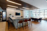This thoughtfully designed amenity space seamlessly blends social, recreational, and professional functionality across a single, open floor. The centerpiece of the lounge is a striking island bar clad in textured tile, topped with light granite and surrounded by a mix of leather and upholstered bar stools — an ideal gathering spot anchored by a wall-mounted TV set against a geometric patterned feature wall. Adjacent to the bar, a fully equipped catering kitchen offers warm wood cabinetry, granite countertops, a sink, stainless steel appliances, and a mounted TV, making it well-suited for events and entertaining. A sun-drenched dining area features a large statement table beneath elegant pendant lighting, surrounded by navy upholstered chairs, with expansive floor-to-ceiling windows framing outdoor views. The spacious open lounge area showcases wide-plank wood-look flooring, a dramatic cluster of gold globe pendants, and a curved cognac leather sectional sofa framed by a slatted wood screen divider. A dedicated game room boasts a billiards table and a bold, colorful mural. Private meeting and conference rooms with glass-panel doors, deep blue carpet, and sheer curtains offer quieter, more focused settings. The overall aesthetic is contemporary and welcoming, defined by natural materials, curated lighting, and an abundance of natural light.