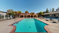 Located at 1800 Stokes St in San Jose, California, this outdoor recreational area is centered around a generously sized swimming pool with vivid turquoise water and a classic brick-trimmed surround. The pool is flanked by a broad concrete deck that provides ample space for lounging, complete with chaise lounge chairs positioned for sunbathing and relaxation. Two elegant gazebos with terracotta-tiled roofs and white structural columns offer shaded gathering spots along the poolside, creating inviting areas to unwind out of the sun. A separate pergola structure on the opposite side of the pool adds additional covered space. The area is enclosed by a sturdy wrought-iron security fence with a gated entry, ensuring a controlled and safe environment for users. Mature deciduous trees surround the perimeter, providing natural shade and a lush, park-like ambiance that softens the hardscape. The surrounding residential buildings are visible beyond the fence line, situating this amenity within a larger community setting. Under the clear California sky, the pool area exudes a bright, open atmosphere well-suited to the warm San Jose climate, offering a well-appointed outdoor retreat for recreation and leisure.