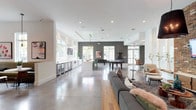 This expansive single-floor amenity space showcases a sophisticated blend of modern and industrial design elements throughout its open, flowing layout. Polished concrete floors extend seamlessly across the entire space, reflecting natural light that pours in through an abundance of large windows framing views of lush greenery outdoors.

The café-style lounge area features a professional coffee station, warm wood cabinetry, built-in banquette seating upholstered in olive green, round café tables with mixed seating, and pendant lighting — creating an inviting spot for casual gatherings. Adjacent to this is a fully equipped demonstration kitchen highlighted by a dramatic chevron-patterned accent wall, a generous white waterfall island with vibrant green barstools, stainless steel appliances, multiple beverage refrigerators, and an industrial-style pendant light fixture.

A billiards and game zone occupies a central area with a pool table set against a dark feature wall, while a dedicated lounge retreat showcases a striking floor-to-ceiling exposed brick accent wall with a mounted flat-screen TV. Plush sectional sofas, eclectic armchairs, layered rugs, and curated artwork complete this comfortable gathering area. Throughout, high ceilings, recessed lighting, and a carefully curated mix of textures contribute to an energetic yet refined ambiance.