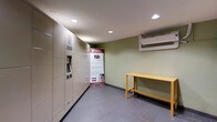 This single-floor package room is designed to streamline parcel management for its users. Dominating the left wall is a full bank of Package Concierge automated lockers finished in a neutral beige tone, equipped with a digital touchscreen kiosk that enables 24/7 self-service package retrieval. A branded informational banner stands nearby, outlining the step-by-step process for registering and collecting deliveries through the system.

The room features sage-green painted walls paired with gray tile flooring, creating a clean and functional aesthetic. Recessed ceiling lighting provides consistent illumination throughout the space. A wall-mounted split air conditioning unit ensures the room remains climate-controlled year-round, an important feature for protecting delivered goods.

Along the far wall, a yellow metal utility bench offers a convenient surface for residents to set down belongings while managing their packages. The overall layout is straightforward and efficiently organized, with ample open floor space allowing for easy movement. The room reflects a focus on practicality and resident convenience, serving as a dedicated, secure hub for package handling within the building.