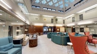 This expansive single-floor space showcases an impressive architectural design centered around a striking coffered skylight ceiling that floods the interior with natural light. The soaring ceiling height creates a grand, open atmosphere, accentuated by a decorative clerestory band of windows and intricate lattice-style molding that encircles the upper perimeter.

The main hall is organized into distinct yet open seating areas. Curved teal sectional sofas form an inviting central gathering space, complemented by warm brown leather chairs and a cream upholstered sofa arranged on a light-toned area rug. Bold coral accent chairs introduce a vibrant contrast to the refined neutral palette. Tall brass floor lamps and fresh floral arrangements add refined finishing touches throughout.

Toward the rear, rich wood-paneled walls frame a secondary dining or meeting area furnished with traditional tables and chairs beneath a classic chandelier. Framed artwork adorns the walls, contributing to the cultured ambiance of the space.

The combination of classical architectural elements—ornate ceiling details, tall columns, and warm wood millwork—alongside contemporary furnishings creates a sophisticated and welcoming environment suited for gathering and collaboration.