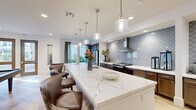 This thoughtfully designed single-floor community space combines modern elegance with functional versatility. The open layout is anchored by a stunning gourmet kitchen featuring an expansive marble-topped island with generous seating, striking arabesque-patterned tile backsplash, dark cabinetry, stainless steel appliances, and a professional-grade range hood. A series of glass pendant lights hang above the island, adding a refined touch to the space.

Adjacent to the kitchen, the main lounge area offers an inviting atmosphere with light wood herringbone-patterned flooring, comfortable seating arrangements, curated artwork, and ample natural light streaming through large windows. The open flow between areas creates a seamless, connected environment ideal for socializing and gathering.

A dedicated beverage station, finished with warm wood cabinetry and the same elegant arabesque tile accent wall, provides a convenient refreshment area near the entrance. Nearby, a carpeted multi-purpose room accommodates flexible seating arrangements around several tables, complemented by a wall-mounted display screen and textured accent wall — well-suited for meetings, events, or casual dining.

Throughout the space, consistent design elements including recessed lighting, clean white walls, and warm wood-toned flooring create a cohesive, welcoming ambiance that balances sophistication with everyday comfort.