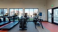 This fitness center is designed to accommodate a wide range of workout styles within a single, thoughtfully organized space. The facility is divided into distinct functional zones, beginning with a dedicated spin studio enclosed by sleek floor-to-ceiling glass partitions with dark frames. The studio features multiple stationary cycling bikes, a large wall-mounted display, full-length mirrors, bold red accent walls, large-scale athletic photography, and an oversized ceiling fan for airflow.

Adjacent to the spin studio, the main strength and cardio area benefits from soaring ceilings that lend an open, airy atmosphere. Large windows along the perimeter flood the space with natural light and offer views of the surrounding outdoor area. The strength zone is outfitted with a comprehensive dumbbell rack, a multi-function cable machine, adjustable benches, and additional resistance equipment. A striking climbing wall mural serves as a focal point at the rear of the room, adding visual energy to the environment.

The cardio zone houses treadmills, ellipticals, and specialty cardio machines, all positioned to take advantage of the expansive window views. Red ceiling panels and linear pendant lighting complement the red, gray, and white modular carpet tiles found throughout. A built-in water station with dual fountains and a storage unit provides practical amenities for users.