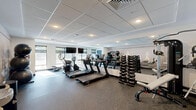 Located at 37 N York Rd in Hatboro, PA, this fitness center offers a well-equipped, single-floor exercise space designed to support a comprehensive workout experience. The room features a modern, clean aesthetic with a drop tile ceiling outfitted with recessed lighting that keeps the space bright and energizing. Large windows along one wall allow ample natural light to fill the room while providing views to the outdoors, complemented by wall-mounted flat-screen televisions for entertainment during workouts. The flooring consists of durable rubber gym matting, practical for both cardio and strength training activities. Cardio equipment includes multiple TRUE-brand treadmills and elliptical machines, each equipped with individual digital displays. A full rack of dumbbells and a cable machine cater to strength training needs, while a TAGfit rack holds an assortment of colorful medicine balls and kettlebells of varying weights for functional fitness routines. Stability balls are also available. A floor-to-ceiling mirror along one wall enhances the sense of openness and allows users to monitor their form. A standout design feature is the large-scale athletic silhouette mural adorning the accent wall, adding a dynamic and motivational atmosphere to the space. A water fountain is conveniently accessible near the entrance, rounding out this fully functional fitness facility.