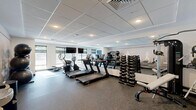 Located at 37 N York Rd in Hatboro, PA, this fitness center offers a well-equipped, single-floor exercise space designed to support a comprehensive workout experience. The room features a modern, clean aesthetic with a drop tile ceiling outfitted with recessed lighting that keeps the space bright and energizing. Large windows along one wall allow ample natural light to fill the room while providing views to the outdoors, complemented by wall-mounted flat-screen televisions for entertainment during workouts. The flooring consists of durable rubber gym matting, practical for both cardio and strength training activities. Cardio equipment includes multiple TRUE-brand treadmills and elliptical machines, each equipped with individual digital displays. A full rack of dumbbells and a cable machine cater to strength training needs, while a TAGfit rack holds an assortment of colorful medicine balls and kettlebells of varying weights for functional fitness routines. Stability balls are also available. A floor-to-ceiling mirror along one wall enhances the sense of openness and allows users to monitor their form. A standout design feature is the large-scale athletic silhouette mural adorning the accent wall, adding a dynamic and motivational atmosphere to the space. A water fountain is conveniently accessible near the entrance, rounding out this fully functional fitness facility.