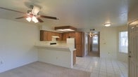 This single-story home offers a practical and open layout designed for comfortable everyday living. The kitchen serves as a central hub of the space, featuring warm wood cabinetry, a tile countertop peninsula that doubles as a breakfast bar, and standard appliances including a stove, refrigerator, and range hood. An overhead skylight-style light fixture adds a bright, airy quality to the kitchen area. The open-concept design allows the kitchen to flow seamlessly into the adjacent living area, which is outfitted with light tile flooring and a ceiling fan with integrated lighting, keeping the space comfortable and well-ventilated. Neutral-toned walls throughout lend a clean, understated aesthetic. A long hallway extends from the main living area, connecting the home's additional rooms and providing a sense of depth and functionality. Natural light filters in from a window visible at the end of the corridor, enhancing the home's bright and welcoming ambiance. The overall design is straightforward and efficient, with a layout that maximizes usability across its single floor.