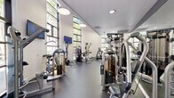 Located at 1201 Race St in San Jose, California, this well-equipped fitness center offers a thoughtfully designed exercise space that accommodates a comprehensive range of workout routines. The room's most striking architectural feature is its soaring double-height ceilings, paired with expansive floor-to-ceiling windows along multiple walls that flood the interior with abundant natural light and offer views of the surrounding greenery, creating an open and energizing atmosphere.

The flooring throughout consists of durable rubberized gym matting, providing both safety and comfort during workouts. Cardio equipment is well-represented, with treadmills, elliptical machines, a stair climber, stationary bikes, and rowing machines positioned to take advantage of the natural light and outdoor views. A dedicated strength training zone includes a multi-station cable machine, a full rack of dumbbells, adjustable benches, medicine balls, TRX suspension trainers, and plyometric boxes for functional fitness training.

A large, bold motivational mural spans one of the upper walls, adding an energetic and urban aesthetic to the space. Full-length mirrors on the strength training walls enhance the sense of openness. Modern pendant lighting complements the natural light, and wall-mounted flat-screen TVs provide entertainment during workouts. The overall design is contemporary, functional, and visually dynamic.