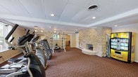 This single-floor amenity facility offers a comprehensive range of shared spaces designed to support both active and relaxed lifestyles. The fitness center is a standout feature, housing an array of cardio machines—including treadmills, ellipticals, and a stationary bike—alongside a multi-station weight rack and bench. Full-length mirrors line one wall, enhancing the sense of space, while a striking floor-to-ceiling stone fireplace serves as a dramatic architectural focal point. Patterned carpeting flows throughout the interior, and a drop ceiling with recessed lighting provides consistent illumination across all areas.

Adjacent to the fitness area, a dedicated business nook offers an L-shaped built-in desk equipped for computer use and printing. A separate dining area accommodates small gatherings around a glass-top table with upholstered chairs, lit by a window overlooking the greenery outside. A functional kitchenette includes a range, double sink, and refrigerator supported by rich wood cabinetry.

Outdoors, a covered deck with a gabled roof and open railings provides shaded seating with direct views of the freeform pool below. The pool area features generous lounge seating and is surrounded by mature trees, creating a private, resort-like atmosphere.